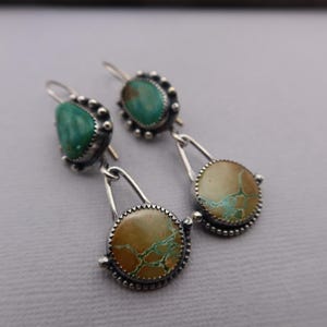 May include: A pair of silver earrings with turquoise and tan-colored stones. The earrings have a teardrop shape with a circular stone at the bottom. The stones are set in a silver bezel with a beaded edge. The earrings are designed for pierced ears.