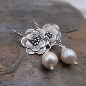 May include: Silver flower earrings with a pearl drop. The earrings feature detailed, openwork flower designs with a cluster of small beads at the center. Each earring has a large, white, teardrop-shaped pearl suspended below.