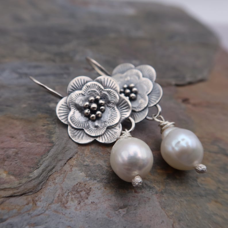 May include: Silver flower earrings with a pearl drop. The earrings feature detailed, openwork flower designs with a cluster of small beads at the center. Each earring has a large, white, teardrop-shaped pearl suspended below.