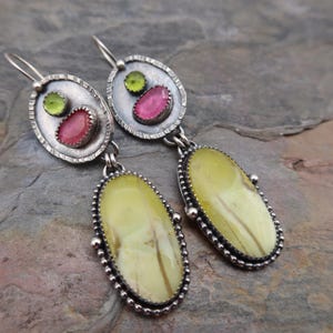Pode incluir: Brincos de prata com um design único. Cada brinco apresenta uma pedra grande, oval, amarelo pálido com veios castanhos, e um oval menor com uma pedra rosa e verde. Os brincos têm uma borda prateada texturizada e fecho de gancho.