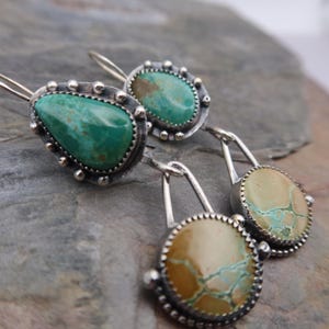 May include: Close-up of handcrafted silver earrings featuring turquoise stones. The earrings have a unique design with a textured silver border and a teardrop-shaped turquoise stone. The other earrings have a round turquoise stone. The earrings are displayed on a gray stone surface.