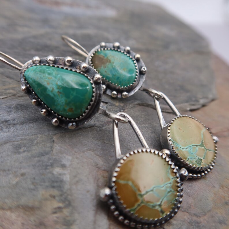 May include: Close-up of handcrafted silver earrings featuring turquoise stones. The earrings have a unique design with a textured silver border and a teardrop-shaped turquoise stone. The other earrings have a round turquoise stone. The earrings are displayed on a gray stone surface.
