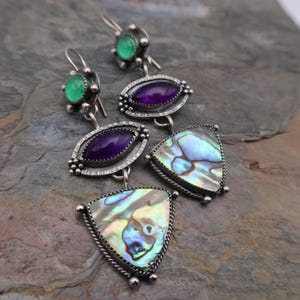 May include: A pair of silver earrings with a green, purple, and iridescent abalone shell design. The earrings feature a small green stone, a larger purple stone, and a triangular abalone shell.