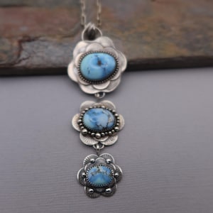 May include: A silver necklace featuring three flower-shaped pendants, each with a light blue oval stone. The stones have a marbled appearance. The necklace is displayed against a gray background.