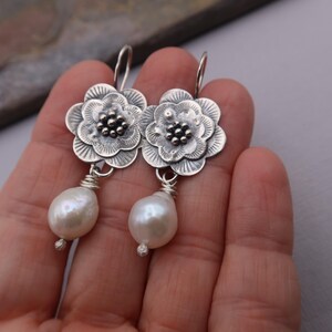 May include: A pair of silver-toned flower earrings with a pearl drop. The earrings feature detailed floral designs with a cluster of small beads at the center. The teardrop-shaped pearls are attached with silver wire.
