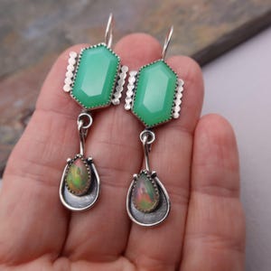 Pode incluir: Um par de brincos pendentes com uma pedra verde hexagonal facetada no topo, ligada por um elo de prata a uma pedra de opala iridescente em forma de lágrima, engastada numa moldura de prata escura com rebordo recortado e gancho.