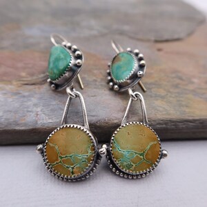 May include: A pair of silver earrings with turquoise and tan-colored stones. The earrings have a unique design, with a teardrop-shaped top and a circular bottom. The stones are set in a silver bezel with a beaded edge.