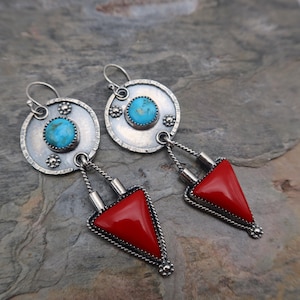 May include: A pair of silver earrings with turquoise and red accents. Each earring features a circular top with a turquoise stone and a red triangular pendant. The earrings have a Southwestern style.