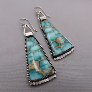 May include: A pair of turquoise and tan gemstone earrings. The earrings have a triangular shape with a silver beaded border and a scalloped edge. The gemstones have a marbled appearance and are suspended from silver ear wires.