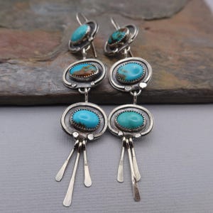 May include: Silver-toned dangle earrings featuring oval turquoise stones. The earrings have a layered design with three turquoise stones per earring, connected by a series of silver circles and dangling silver accents.
