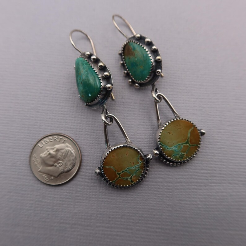 May include: A pair of silver earrings with turquoise and tan-colored stones. The earrings have a unique design with a teardrop-shaped turquoise stone at the top and a circular tan stone at the bottom. The earrings are approximately 2 inches long.