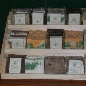 May include: A wooden display stand with three shelves, each holding a variety of handmade soap bars. The soaps are in various colors and patterns, including green, brown, and white. The soaps are labeled with names like "Peppermint Patty" and "The Christmas Cookie".