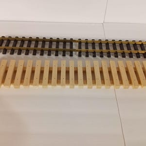 May include: A wooden track bed for model trains, with a set of metal track pieces on top. The track bed is made of light brown wood and has a series of evenly spaced holes. The track pieces are made of dark brown metal and have a gold-colored finish.