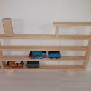 May include: Wooden train shelf with three shelves and a top shelf. The shelf is painted in a natural wood color. There are three toy trains on the shelves, one blue, one yellow, and one gray.