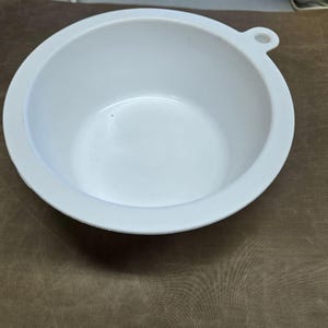 May include: A white, round bowl with a handle on the side. The bowl has a smooth, glossy finish and a simple, minimalist design. The bowl is likely made of plastic or ceramic and is suitable for various uses.