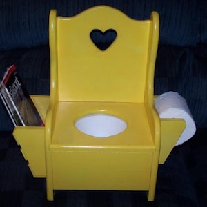 May include: A yellow wooden potty chair with a heart-shaped cutout in the back. The chair has a white toilet seat and a roll of toilet paper on the side. There is a book in the side pocket.