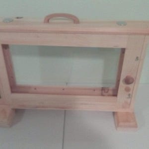 May include: A light wood frame with a rectangular opening and a handle on top. The frame has a simple design and is made of unfinished wood.