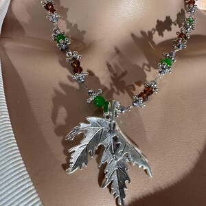 Peut inclure: Un collier composé d'un grand pendentif en forme de feuille d'érable argentée, suspendu à une chaîne de perles alternées vertes et ambrées, entrecoupées de petites entretoises argentées et d'accents métalliques au design botanique.
