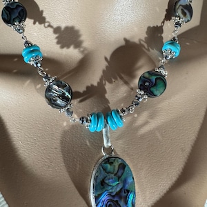 May include: A necklace featuring a pendant with iridescent blue and green abalone shell. The necklace has turquoise beads and silver-toned accents. The design includes a mix of round and cylindrical beads, creating a unique jewelry piece.