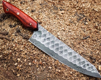 May include: A Damascus steel chef's knife with a patterned blade and a reddish-brown wooden handle. The knife is lying on a bed of wood shavings, showcasing its craftsmanship and design.