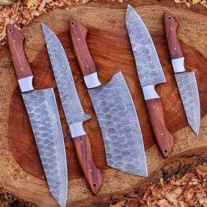 May include: A set of six kitchen knives with patterned steel blades and dark wood handles. The knives are arranged on a wooden surface, showcasing their various sizes and shapes, including a cleaver. The handles are riveted with brass pins.