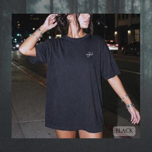 May include: A black short-sleeve t-shirt featuring a small white script text on the left chest that reads angel with a halo graphic above it. The garment has a crew neckline and a relaxed fit, shown in a dark outdoor setting at night.