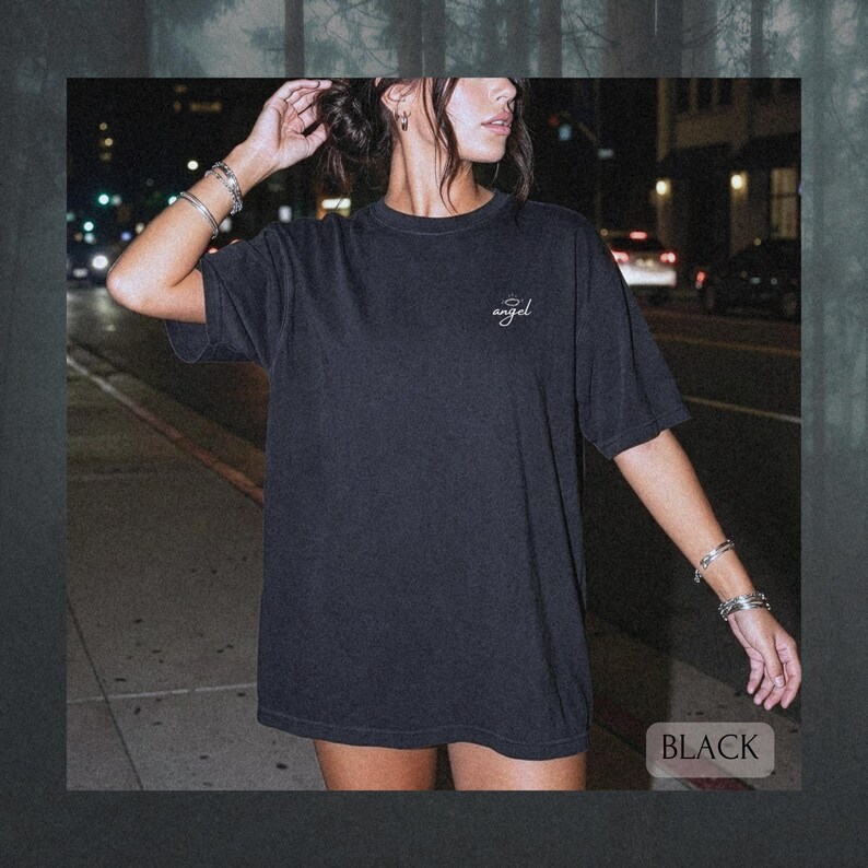 May include: A black short-sleeve t-shirt featuring a small white script text on the left chest that reads angel with a halo graphic above it. The garment has a crew neckline and a relaxed fit, shown in a dark outdoor setting at night.