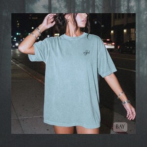 May include: A sage green short-sleeve t-shirt with a crew neckline and a relaxed fit. The front features the word "angel" printed in a small black cursive script on the upper left chest, accompanied by a simple line-drawn halo graphic above it.