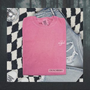 May include: A pink short-sleeved t-shirt with a crew neckline. The shirt features the word angel in white cursive script with a halo above it on the upper right chest and the word CRUNCHBERRY in a white rectangular box at the bottom right.