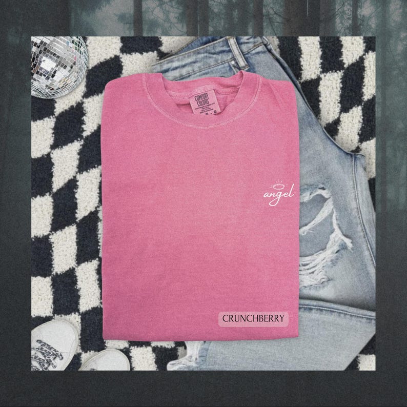 May include: A pink short-sleeved t-shirt with a crew neckline. The shirt features the word angel in white cursive script with a halo above it on the upper right chest and the word CRUNCHBERRY in a white rectangular box at the bottom right.