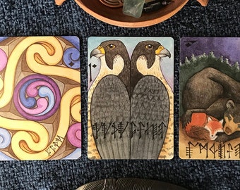 May include: An assortment of tarot cards with illustrations of birds, a bear and fox, and abstract designs. A wooden bowl holds crystals and charms. A silver feather rests on a blue textured surface. The cards have runic inscriptions.