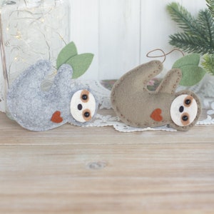 May include: Two felt sloth ornaments, one gray and one brown, with stitched details and red hearts. The ornaments are hanging from a white background.