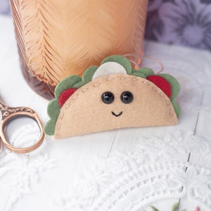 May include: A handmade felt taco ornament with a smiling face, black button eyes, and red, green, and white fillings. The taco is tan with a stitched outline. A pair of decorative scissors and a glass vase are in the background.