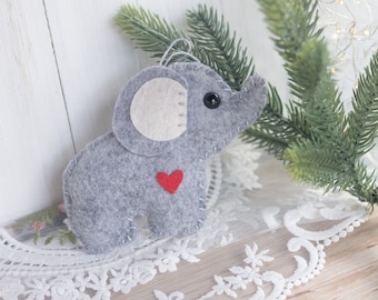 May include: A gray felt elephant ornament with a white felt ear and a red felt heart on its chest. The ornament is hanging from a green pine branch.