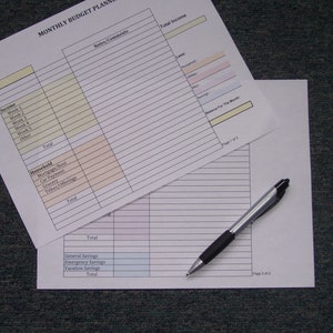 May include: A two-page printable budget planner with sections for income, household expenses, and savings. The planner has lines for writing in amounts and notes. The planner is on a gray surface with a silver pen.