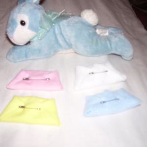 May include: Four fabric squares with safety pins, in pink, yellow, white, and blue. The squares are arranged in a row on a white surface. A blue plush bunny toy is in the background.
