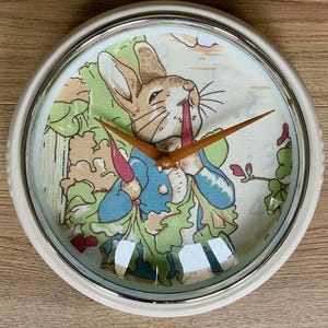 May include: A round wall clock with a white frame and a glass cover. The clock face features an illustration of a brown rabbit wearing a blue jacket, eating a carrot, surrounded by green leafy vegetables. It has two orange clock hands.