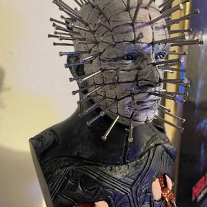 May include: A bust of a horror movie character with a white, textured face covered in nails. The character has a black leather jacket with a red and black strap.