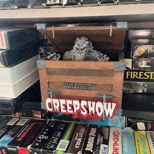 May include: A wooden box with a monster popping out of it. The box has a sign that says "Creepshow" and "Arctic Expedition June 18, 1834". The box is surrounded by VHS tapes.