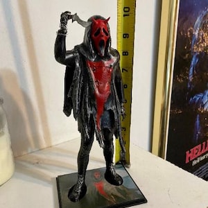 May include: A black and red 3D printed figurine of a masked figure holding a knife. The figure is wearing a black hooded robe with a red lining and a red mask with a scream face. The figure is standing on a black base with a printed image.