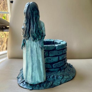 Może przedstawiać: Tealowa figurka kobiety z długimi włosami stojąca obok kamiennego studni. Figurka jest wykonana z fakturowanego materiału i ma postarzony wygląd.