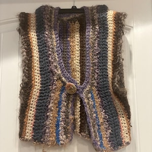 Puede incluir: Un chaleco de ganchillo hecho a mano, multicolor, con cierre de botón. El chaleco presenta rayas verticales en tonos marrón, beige, azul y morado. El hilo texturizado crea un aspecto bohemio y acogedor. El chaleco se muestra en una percha negra contra una puerta blanca.