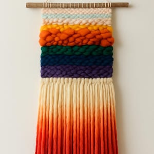 May include: A colorful woven wall hanging with a rainbow design. The top section features horizontal bands of pastel pink, light blue, yellow, orange, green, dark blue, and purple. The bottom section has long, vertical strands that fade from cream to orange.