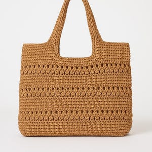 Könnte beinhalten: Eine hellbraune Häkeltasche mit zwei Griffen. Die Tasche hat eine rechteckige Form mit einem strukturierten, offen gewebten Design. Die Griffe sind ebenfalls gehäkelt und oben an der Tasche befestigt.