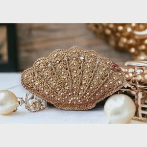 May include: A handcrafted, seashell-shaped clutch in a warm, golden-brown hue. The crochet design features intricate openwork and is embellished with small, white beads. The clutch is a unique accessory, perfect for beach-themed events or adding a touch of coastal charm.