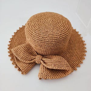 Peut inclure: Chapeau de paille tissé de couleur marron clair, avec un large bord et un grand nœud. Le bord du chapeau est festonné. Le nœud est situé à l'avant du chapeau, idéal pour l'été.