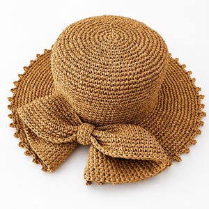 Puede incluir: Un sombrero de paja trenzada de color tostado con una copa redondeada y un ala ancha con borde festoneado. Un lazo grande de paja trenzada a juego está sujeto en la parte delantera del sombrero, añadiendo textura y detalle a este accesorio.