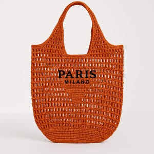 Op de afbeelding: Een oranje gehaakte tote bag met een grote handgreep. De tas heeft de woorden "PARIS MILANO" in zwarte blokletters. De tas heeft een geweven, open ontwerp.