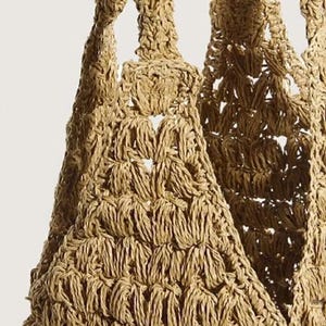 May include: A tan-colored tote bag crafted with a textured, open-weave crochet pattern. The bag features thick, integrated shoulder straps that transition into a triangular gusset design, showcasing a consistent, knotted fiber construction throughout.