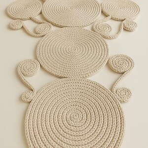 May include: A decorative wall hanging composed of beige rope. The design features multiple circular and spiral elements, connected by curved rope segments. The rope is tightly woven, creating a textured surface. The overall shape is abstract and artistic.
